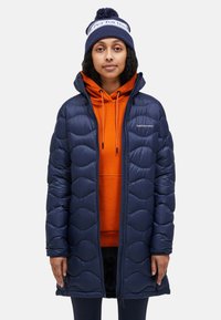 Marine blauwe pufferjas met gewatteerd ontwerp, rits aan de voorkant en capuchon. Draagt over een oranje hoodie, zichtbare trekkoorden en zachte textuur. Gebreide muts met pompon.