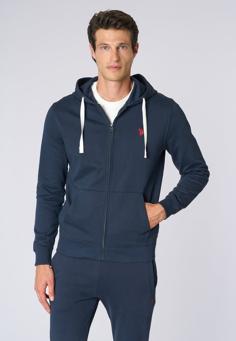 Felpa con zip di colore blu navy in tessuto morbido, dotata di tasca frontale, cappuccio regolabile con cordino e dettaglio logo rosso sul petto.