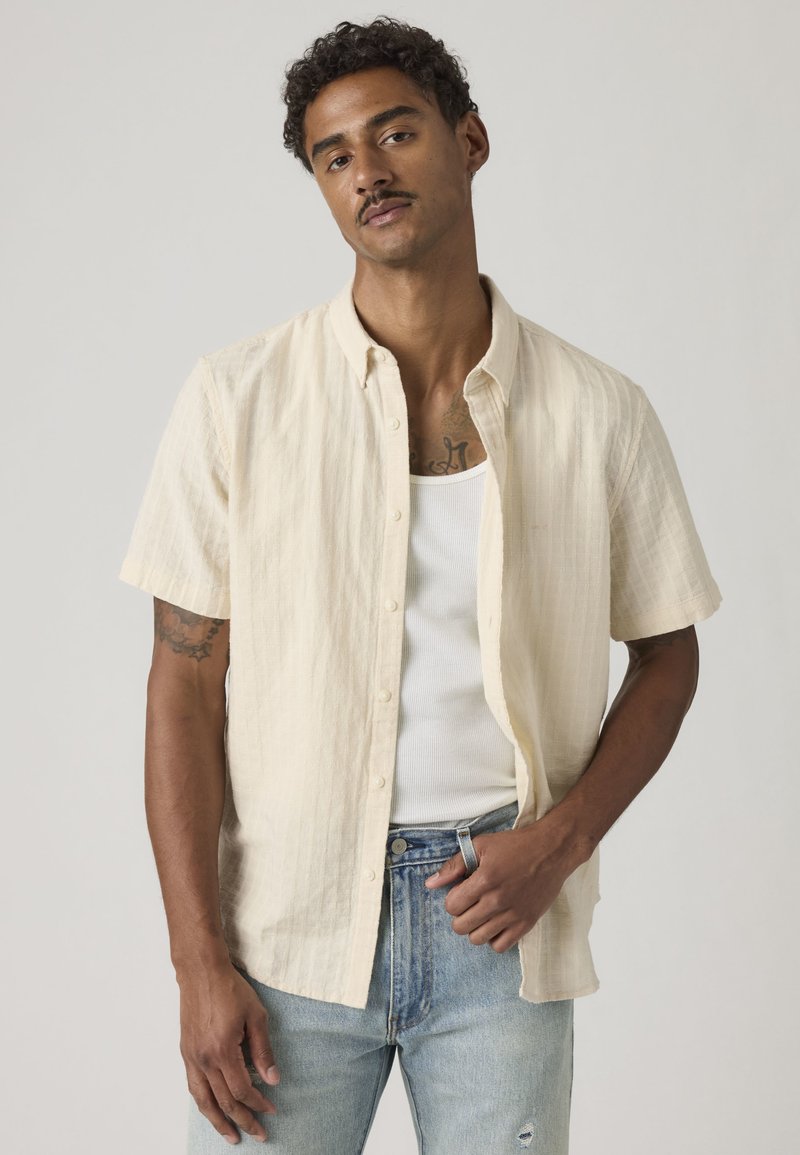 Homme aux cheveux bouclés portant une chemise beige à manches courtes par-dessus un débardeur blanc et un jean bleu clair, se tenant devant un fond uni.