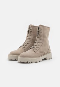 Beige nahkaiset nilkkasaappaat, joissa on pyöreät varpaat, nauhakiinnitys edessä ja paksut, karkeat kumipohjat. Sisältää teksturoidut yksityiskohdat ja vahvistetut saumat.