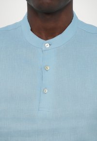 Lys blå lin skjorte med mandarin krage, tre knapper og subtil tekstur. Glatt stoff med minimal sømdetalj.
