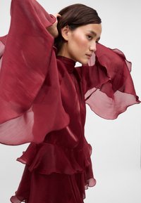 Burgunderfarget kjole med gjennomsiktige, vide bell-sleeves og rynkede lag i kanten. Glatt tekstur med høy hals og lagdelte designdetaljer.