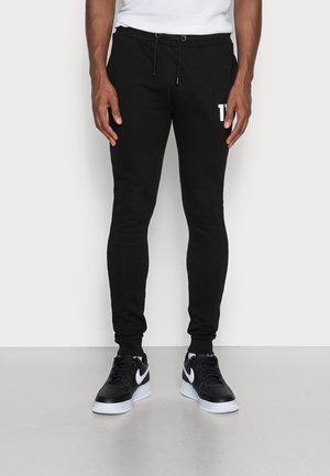 Homme portant un jogger noir slim avec un cordon blanc et des baskets Nike noires et blanches, debout sur un sol gris.