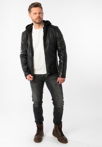 Giacca di pelle nera con cappuccio nero attaccato, chiusura con zip sul davanti, spalle a coste, abbinata a jeans grigi e stivali marroni con lacci.