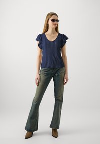 Mørkeblå topp med korte volangerm, v-hals og et strukturert mønster, kombinert med lys bleket vide jeans og brune spisse støvler.