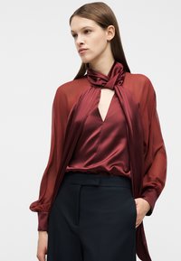 Γυναίκα που φοράει μπλούζα από σατέν μπορντό με λαιμόκοψη φουλαρί και διαφανή μακριά μανίκια, συνδυασμένη με μαύρο παντελόνι ψηλόμεσο, με το χέρι στην τσέπη, κοιτάζοντας προς τα αριστερά.