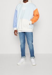 Felpa con blocchi di colore caratterizzata da blu chiaro, arancione e bianco, con tasca a marsupio, abbinata a jeans blu chiaro e sneakers bianche.