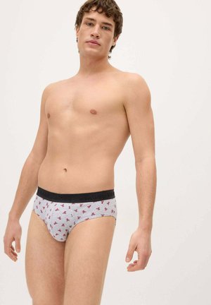 Hombre joven de pie con calzoncillos grises con pequeño estampado floral rojo y cintura negra sobre un fondo claro y liso.