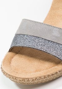 Gros plan d'une sandale beige avec une semelle intérieure douce et deux brides : une grise mate et une argentée pailletée texturée.