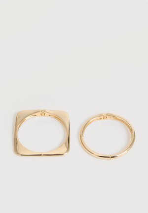 Deux bracelets en métal doré côte à côte, l'un de forme carrée et l'autre circulaire, tous deux avec une finition lisse et polie sur fond blanc.
