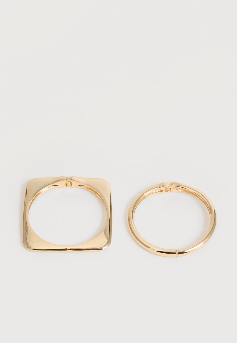 Δύο βραχιόλια από μέταλλο σε χρυσαφί απόχρωση δίπλα-δίπλα, ένα τετράγωνο και ένα κυκλικό, και τα δύο με λείο, γυαλιστερό φινίρισμα σε λευκό φόντο.