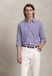 Cămașă lungă cu mâneci, de culoare purpurie deschisă, cu nasturi, purtată cu pantaloni albi și o curea maro. Cămașa are un logo subtil pe piept.