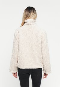 Pull en polaire beige avec un col haut, texture douce et coupe décontractée. Présente des manches longues et un ourlet arrondi à l'arrière.