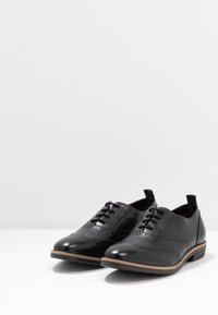 Chaussures en cuir noir avec un design brogue, présentant des accents perforés, un système de laçage et un talon plat avec une semelle marron clair.
