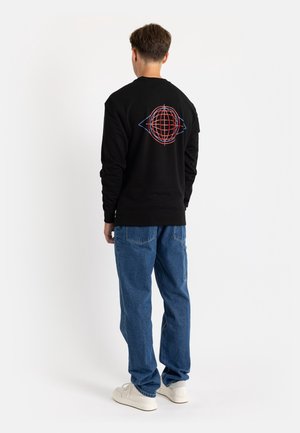 Ung mand iført sort sweatshirt med rød og blå globusgrafik på ryggen, blå jeans og hvide sneakers, set bagfra.
