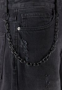 Un bolso de mezclilla negra que presenta un parche desgastado y una cadena de metal negro unida a un lazo. La cadena tiene eslabones grandes con un acabado brillante.