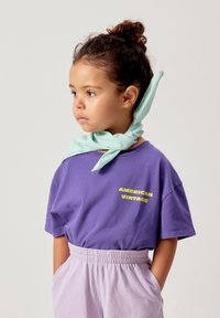 Jeune fille aux cheveux bouclés attachés, portant un t-shirt violet "American Vintage", un pantalon violet clair et une écharpe vert menthe autour du cou.