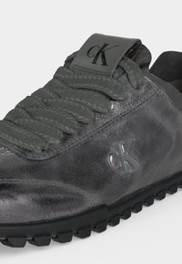 Baskets en cuir gris foncé avec des lacets gris épais texturés, semelle en caoutchouc noire, et logo "cK" embossé sur le côté et la languette.