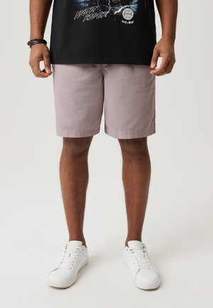 Jasnoróżowe szorty z bawełny, do kolan, z bocznymi kieszeniami i prostym wykończeniem, w połączeniu z białymi sneakersami. Na górze czarny t-shirt z grafiką.