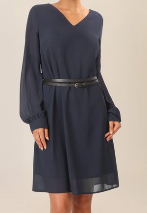 Femme portant une robe bleu marine à manches longues et col en V avec une ceinture noire à double sangle à la taille, sur un fond uni.