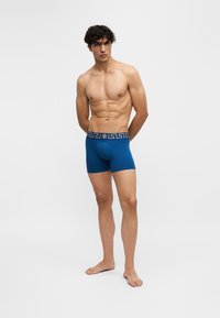 Homme torse nu aux cheveux bouclés foncés, debout pieds nus, portant un boxer bleu avec une ceinture décorative sur un fond blanc uni.