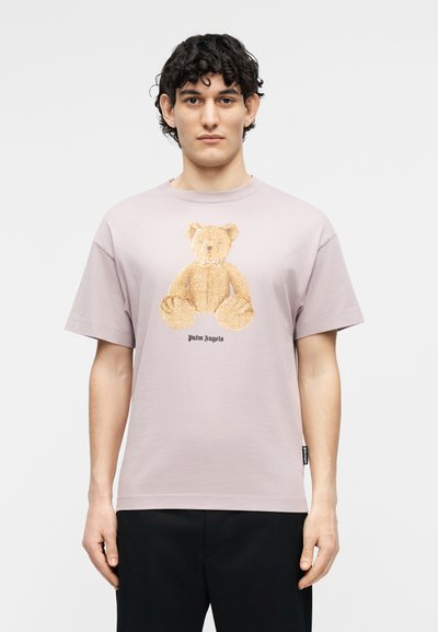 Νεαρός με σγουρά μαλλιά που φοράει ένα ανοιχτό μωβ μπλουζάκι με ένα μεγάλο γραφικό αρκουδάκι και το κείμενο "Palm Angels", στέκεται μπροστά σε ένα απλό φόντο.