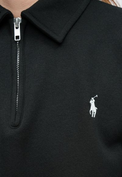 Svart zip-up polo laget av strikket stoff med en krage. Har en hvit brodert logo av en rytter med en kølle på venstre side.