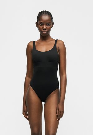 Γυναίκα με πλεξούδες, φορώντας μαύρο ολόσωμο μαγιό χωρίς μανίκια, στέκεται μπροστά σε ένα απλό ανοιχτό γκρι φόντο, κοιτώντας ευθεία μπροστά.