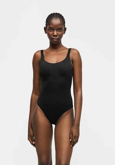Femme aux cheveux tressés portant un maillot de bain une pièce noir sans manches, debout devant un fond gris clair uni, face à l'avant.