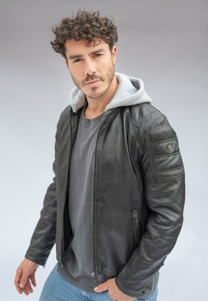 Mężczyzna o kręconych włosach, ubrany w czarną skórzaną kurtkę na szarym kapturze i koszulce, stojący na tle jednolitego jasnoszarego tła.