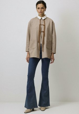 Kobieta stoi, ubrana w beżowy kardigan z otwartym przodem i skórzanymi troczkami, na białej koszuli z kołnierzykiem, w połączeniu z niebieskimi rozszerzanymi dżinsami i beżowymi szpilkami.