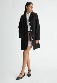 Cappotto nero con bottoni, maniche lunghe e tasche laterali. Indossato sopra una camicia bianca e una gonna a quadri rosa; abbinato a ballerine nere.