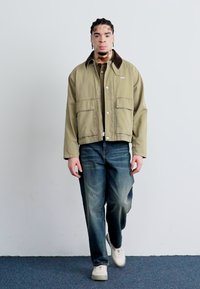 Olivengroene bomberjack met een bruine kraag, twee voorzakken, gelaagd over een textuur gebreid top en gecombineerd met licht vervaagde blauwe jeans.