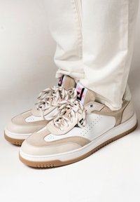 Beige and white sneakers with suede and leather textures, featuring perforated detailing, patterned laces, and gum rubber soles.
