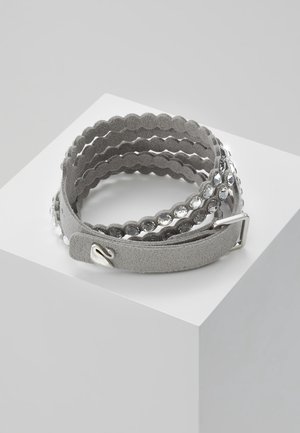 Grå ruskind wrap-armbånd med bølgede kanter og klare runde rhinsten, viklet rundt på en hvid minimalistisk piedestal.