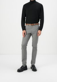 Muž oblečený v černém roláku, šedých kalhotách s hnědým opaskem, šedých ponožkách a černých kožených mokasínech stojící proti bílé background.