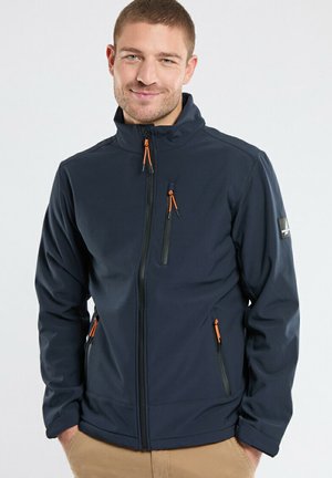 Homme portant une veste zip-up bleu marine avec des tirettes de fermeture éclair orange et un pantalon beige, souriant, mains dans les poches, fond neutre.
