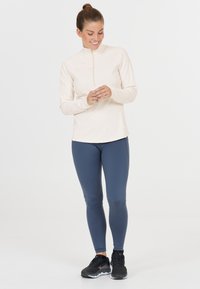 Helles beige Langarmoberteil mit halben Reißverschluss, kombiniert mit dunkelblauen Leggings. Schwarze Sportschuhe vervollständigen das Outfit. Glatte Stofftextur.