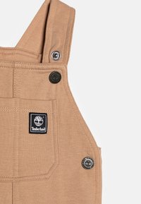 Salopette in cotone color cammello con spalline regolabili, tasca sul petto e dettagli in metallo. Presenta un patch del logo Timberland in nero.