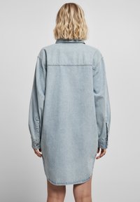 Valkoinen sininen farkkukangasmekko pitkällä hihoilla, kauluksella ja pyöristetty hem. Varustettu napitetuilla hihansuilla ja rennolla leikilla.