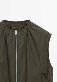 Veste sans manches olive foncée avec encolure ronde froncée et fermeture zippée argentée centrale sur fond blanc.