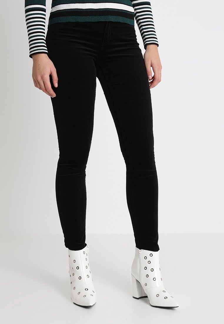 Žena oblečená v černých skinny kalhotách, zeleno-bílém pruhovaném svetr a bílých kotníkových botách s kulatými kovovými akcenty.