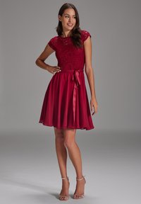 Robe rouge à manches cap avec une jupe en chiffon évasée, lien en satin à la taille et une texture lisse. L'ourlet tombe au-dessus du genou.