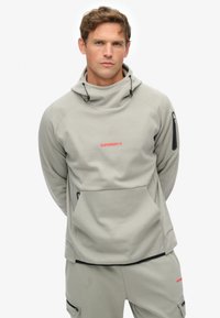 Sudadera de color gris claro hecha de un tejido suave, con un cuello alto, bolsillo frontal, logo rojo y detalles en negro en las mangas.
