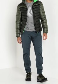 Mies, joka on pukeutunut vihreään ja mustaan toppatakkiin, harmaaseen logolliseen t-paitaan, tummansiniset juoksuhousut ja vihre ät vaelluskenkä, seisoo valkoista seinää vasten.