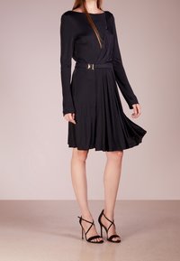 Robe noire à manches longues avec un corsage ajusté et une jupe évasée. Présente une ceinture à la taille avec une boucle métallique et un tissu lisse et fluide.