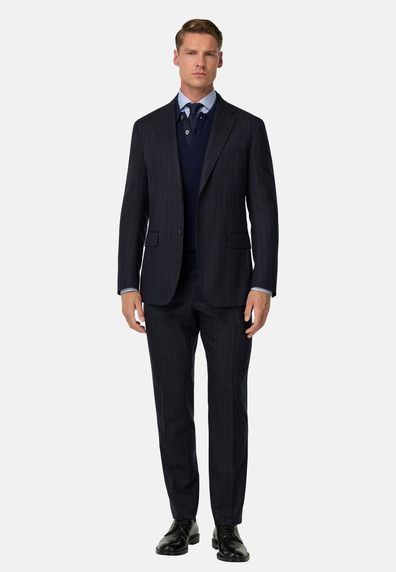 Jūras spēku zils pinstripe uzvalks ar vienu pogu, pielāgota silueta, apvienots ar jūras spēku zila džemperi un baltu kreklu. Melni uzvalka apavi pabeidz izskatu.