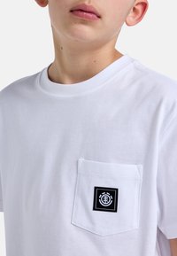 Bílý bavlněný tričko s kulatým výstřihem a levou kapsou na prsou, na které je černá nášivka s logem. Jednoduchý design s jemnou strukturou.