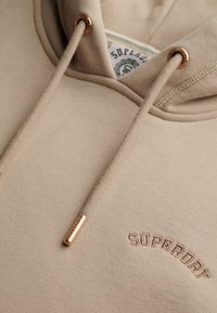 Beige hoodie met een kangoeroezak, geborduurd "SUPERDRY" logo, metalen oogjes en koorden met koperen accenten. Soepele textuur.