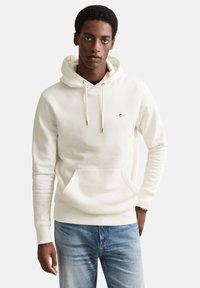 Sweat à capuche crème avec une poche avant, une capuche à cordon et un petit logo. Il présente une texture douce et des poignets côtelés. Associé à un jean bleu clair.